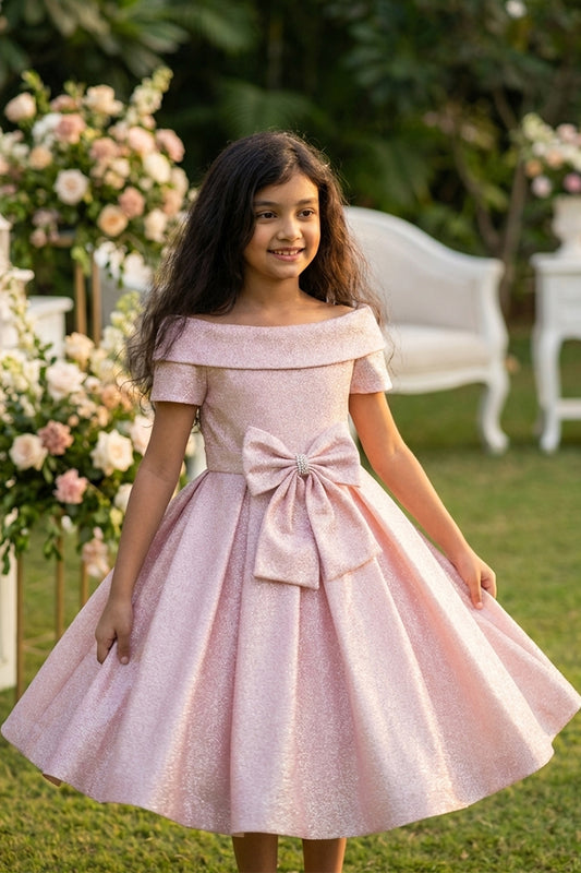 Young girl in a pink off-shoulder dress with a bow standing outdoors.