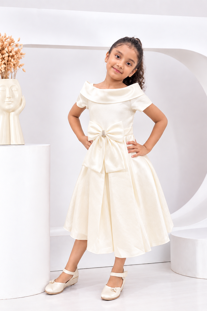 A young girl models an off-the-shoulder cream satin dress with a large bow at the waist, paired with matching cream shoes, in a modern white studio setting.