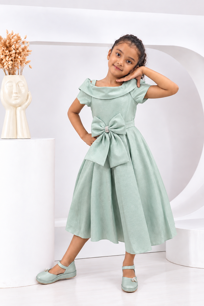 A young girl models a mint green formal dress with a large bow at the waist, styled with matching mint green shoes in a clean, modern white studio setting.