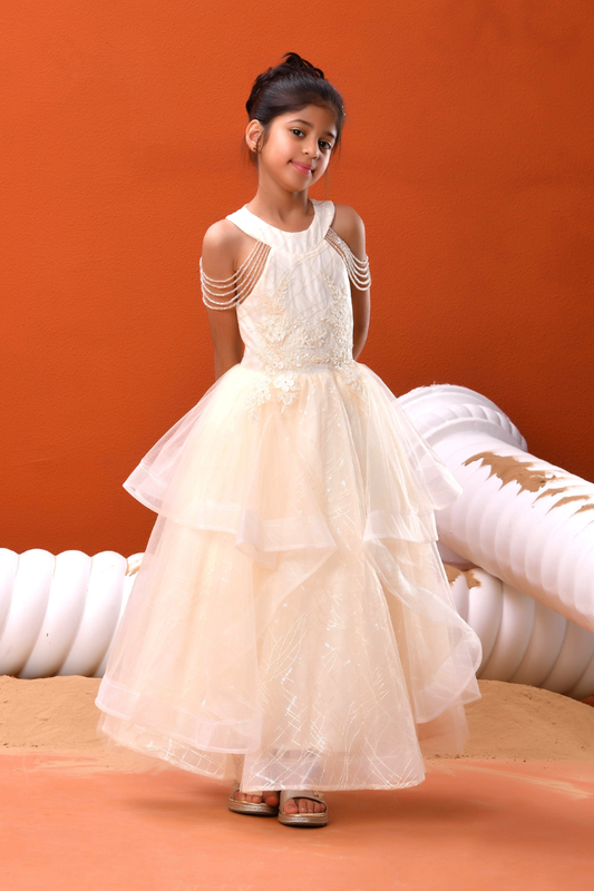 A young girl modeling a formal cream tulle dress with sheer shoulder straps and delicate bead details, set against a warm orange background with white sculptural props.