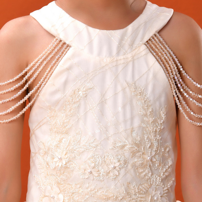 A close-up of a child wearing a cream or ivory formal dress with floral lace applique and pearl-bead draped shoulder straps, set against a solid orange background.