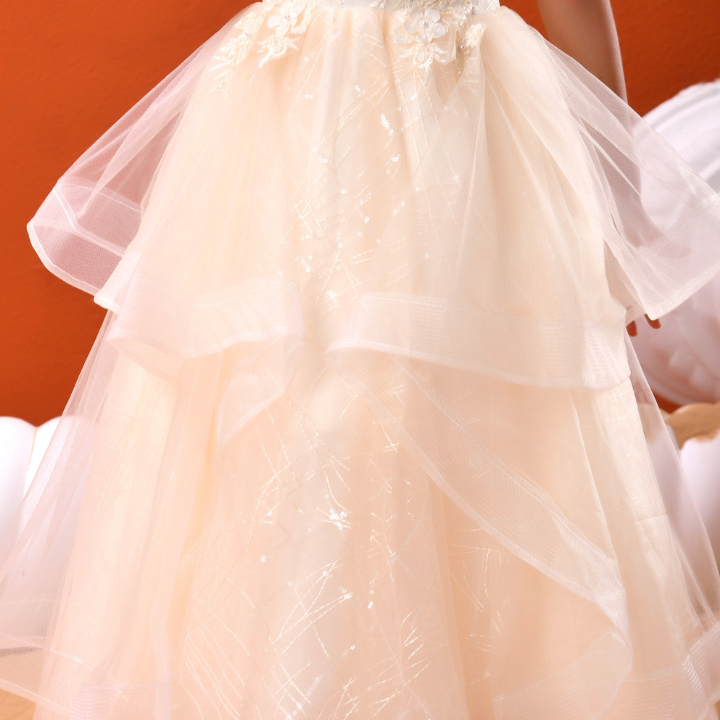 A close-up of a child's feet wearing a long, layered tulle dress in a warm cream or ivory shade, standing against a solid orange background.