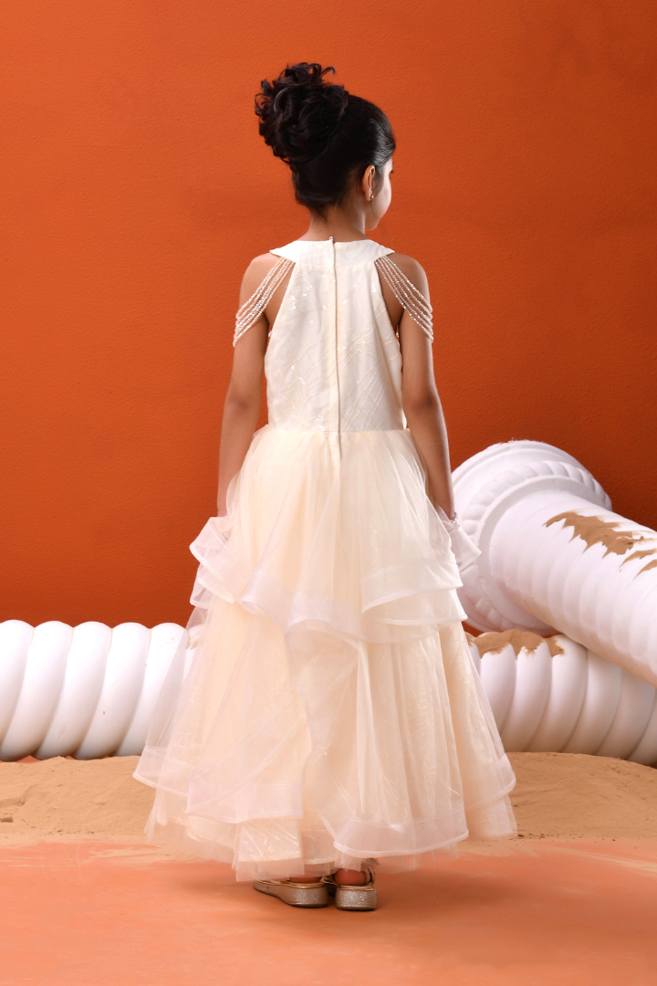 A young girl viewed from behind wearing a sleeveless cream tulle dress with sheer shoulder straps, standing against a solid orange background.