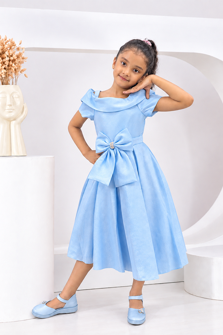 A young girl models a light blue dress with ruffled sleeves, a matching waist tie with a large bow, and light blue flat shoes in a clean, modern white studio setting.