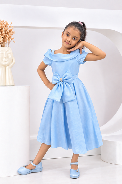 A young girl models a light blue dress with ruffled sleeves, a matching waist tie with a large bow, and light blue flat shoes in a clean, modern white studio setting.