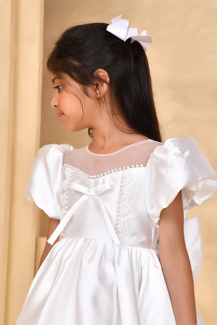 Young girl wearing a white dress with puffed sleeves and a bow detail, against a beige background.