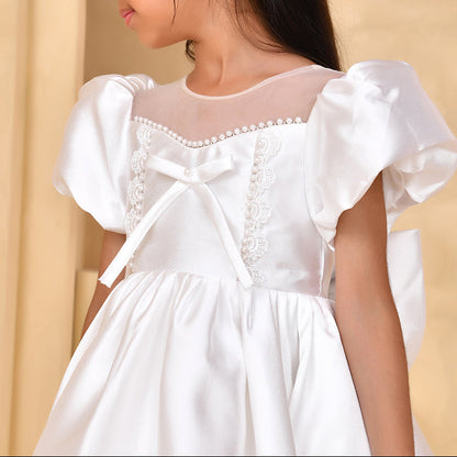 Young girl wearing a white dress with puffed sleeves and a bow detail, against a beige background.
